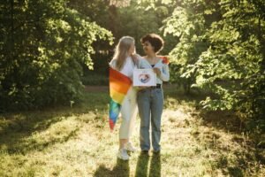 An interracial lesbian couple celebrating pride outdoors, wrapped in a rainbow flag, smiling lovingly at each other.