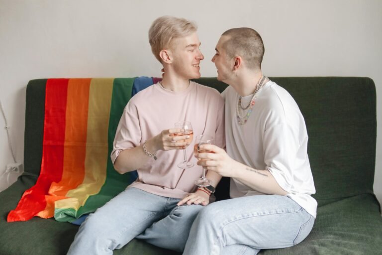 A joyful LGBT couple sitting on a sofa with a rainbow flag, sharing drinks and affection indoors.